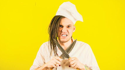Young serious man dressed as chef with knives on yellow background. Male cook in white uniform sharpening crossed knives. Concept of preparing to make food.