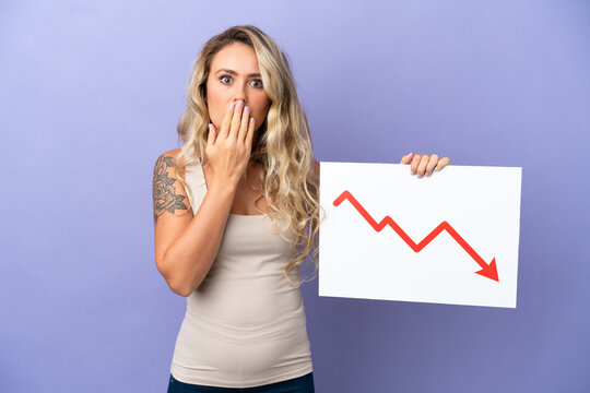 Young Brazilian woman isolated on purple background holding a sign with a decreasing statistics arrow symbol with surprised expression