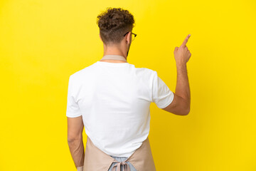 Restaurant waiter blonde man isolated on yellow background pointing back with the index finger