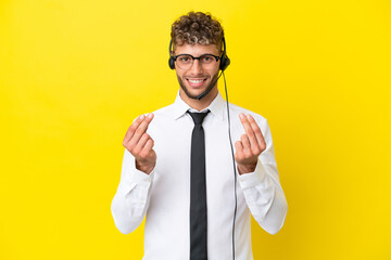 Telemarketer blonde man working with a headset isolated on yellow background making money gesture