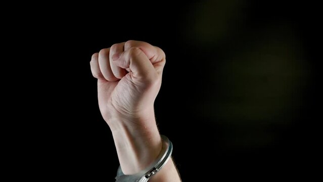 rised fist of caucasian hand shakled in siver handcuffs against the black background.
