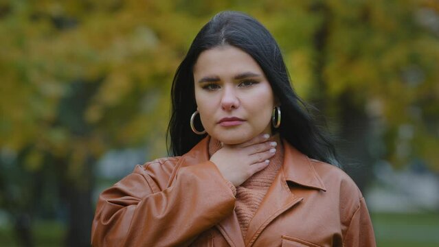 Portrait young sad hispanic woman standing outdoors feeling sore throat suffering from painful symptoms flu infection holding hand on neck chronic tonsillitis spasm allergy frustrated with discomfort