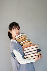 Portrait of puzzled Asian student girl in knitted vest carrying big stack of books against isolated background