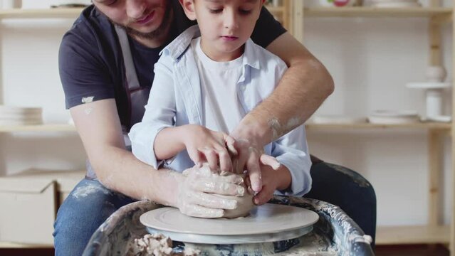 Close up of man and his son are making pot from clay on throwing wheel in studio busy with creative activity. People and family hobby concept.