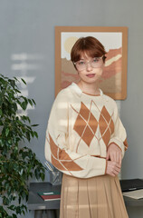 Portrait of confident attractive student girl in old-fashioned look standing against table and picture on wall