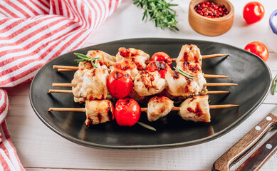 Grilled chicken skewers with cherry tomatoes on a black plate. White wooden table.