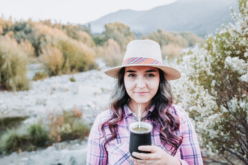 Young caucasian woman with a hat and wearing a pale pink blouse, drinking mate in a natural space with an autumnal mood.