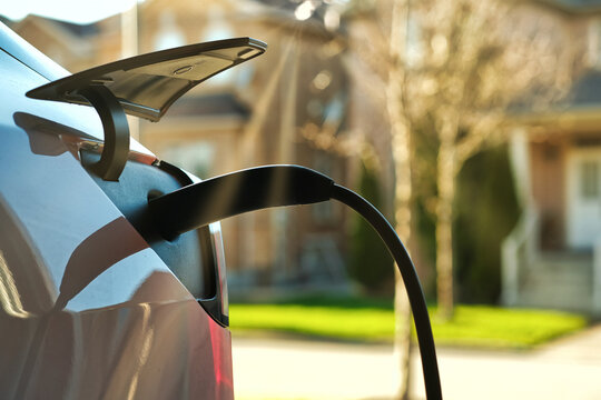 EV Electric Car Plugged Charging At Residential Home Under Sunset Light Against Blurred Street And House Around Background. Close Up.