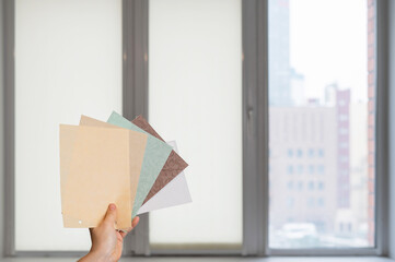 Woman holding fabric samples of roller blinds against window background. 