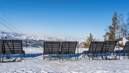 Wooden benches stand in a row on the edge of a snow-covered plateau. Ahead, against the blue sky -...