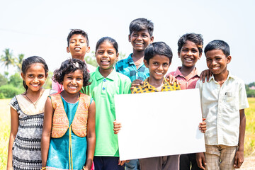 Group of happy smiling Indian village children with one kid holding empty sign board or placard by...