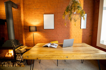 Liiving room interior with a wooden dining table, burning fireplace and brick wall background at home. Laptop and magazine on the table top