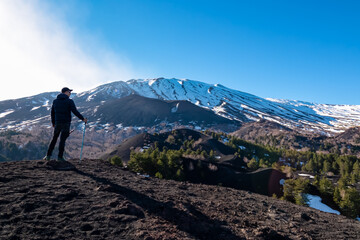 Obraz premium Man with hiking pole on volcanic landscape of volcano mount Etna, in Sicily, Italy, Europe. Pine and white birch trees growing on solidified lava, ash and pumice. Slopes of crater covered with snow