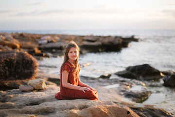 A European teenage girl in a dress sits on stones on the seashore, peace and quiet, a lonely child looks at the sea