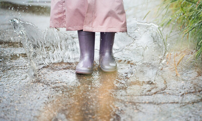 Little girl in pink waterproof raincoat, purple rubber boots funny jumps through puddles on street road in rainy day weather. Spring, autumn. Children's fun after rain. Outdoors recreation, activity