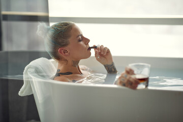 A young beautiful tattooed girl enjoying a cigar and a drink while having a warm bath in the...