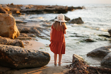 child in a dress and a hat walks barefoot along the seashore, a girl walks along the water's edge, rest and tranquility on the sea coast, rocks and red stones