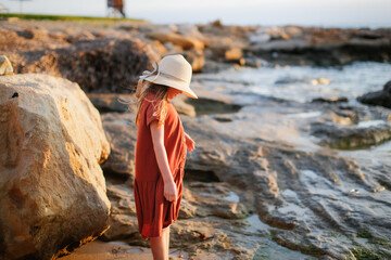 child in a dress and a hat walks barefoot along the seashore, a girl walks along the water's edge,...