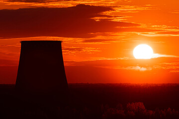 The silhouettes of the cooling tower of thermal power station among the forest on the background of scarlet sunrise or sunset. The cloudy sky cloured in red, orange, rose, scarlet and crimson