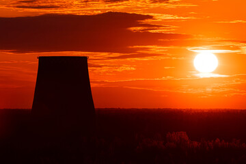 The silhouettes of the cooling tower of thermal power station among the forest on the background of scarlet sunrise or sunset. The cloudy sky cloured in red, orange, rose, scarlet and crimson