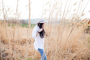 Naklejka na ściany i meble Young brunette attractive woman with long hair, wearing white blouse, outdoors, standing in a field in wind, on blue sky background. 