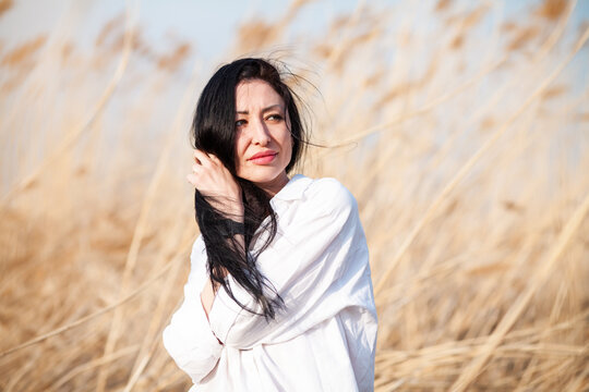 Young brunette attractive woman with long hair, wearing white blouse, outdoors, standing in a field in wind, on blue sky background. 