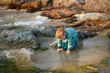 Toddler girl in a dress plays with water on the seashore at sunset, sand and stones, lifestyle