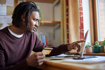 Male University Or College Student With Credit Card Looking At Laptop Worried About Debt At Desk In Room