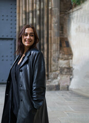 three quarter length shot of a smiling woman of latin features dressed in black with hands in pockets standing on a church's square looking away.
