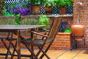 Fototapeta na wymiar Heavy rain falling on the garden furniture made of teak wood and flowering plants.
