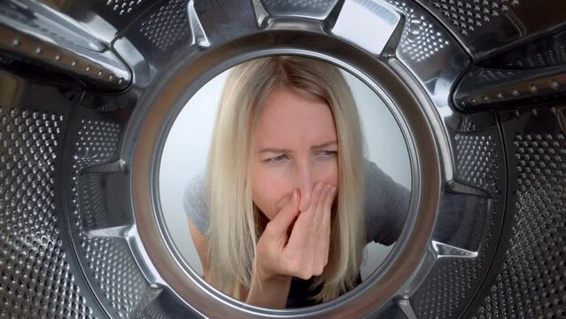 Smelly washing machine and laundry room. Young blonde woman looks inside washing machine, smells stench and plugs her nose. View from inside drum at wide angle. odor elimination. Young hostess.