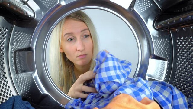 Young Caucasian housewife looks into drum of washing machine, takes out shirt with hole on it. Washing machine ruined clothes, tore them up. Woman is very surprised and upset. Call wizard, warranty