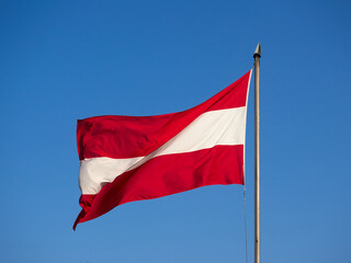 Naklejka na ściany i meble The flag of Austria hanging on a mast. A flag fluttering in the wind against a blue sky.