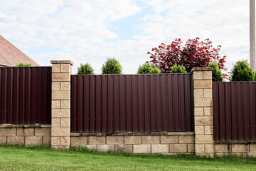 Brown metal profile fence with block posts. Incline construction. Corrugated surface. Security. Private property fencing. Opaque hedge. Outdoor house exterior. Side view. Urban or industrial style