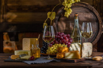 Cheese and wine tasting still life. Glass of wine, bottle of wine, grape in front of the rustic wooden barrels for beverage in a dark wine cellar. Nuts, honey, dipper. Empty copy space, mock up