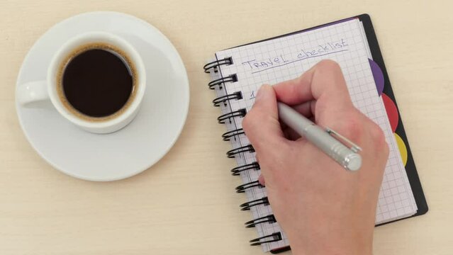 Male hand writes with a pen in a notebook on the table near a white cup of espresso coffee on a saucer travel checklist