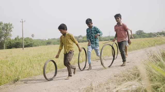 Indian village kids playing with tyre wheel rolling near paddy field rural street - concept of entertainment, holidays and leisure activities.