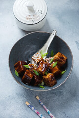 Dark-grey bowl with fried tofu cubes in teriyaki sauce, studio shot on a light-blue stone background
