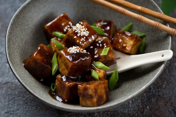 Closeup of fried teriyaki tofu with scallions and white sesame served in a grey bowl, selective focus