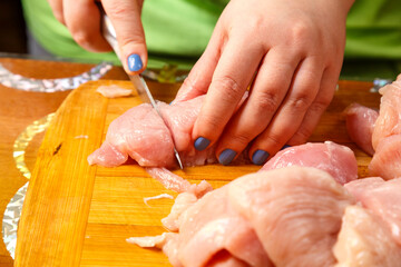 A woman's hands cut a chicken fillet with a large knife on a wooden board in the kitchen.