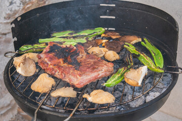Black round metal barbecue with ribs and vegetables already cooked on the grill. Round metal container for cooking barbecues with a grill placed on top full of fresh meat, mushrooms and green peppers.