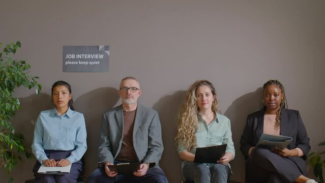 Zoom in group portrait of multiethnic job candidates sitting with clipboards in office corridor and posing for camera while waiting for interview