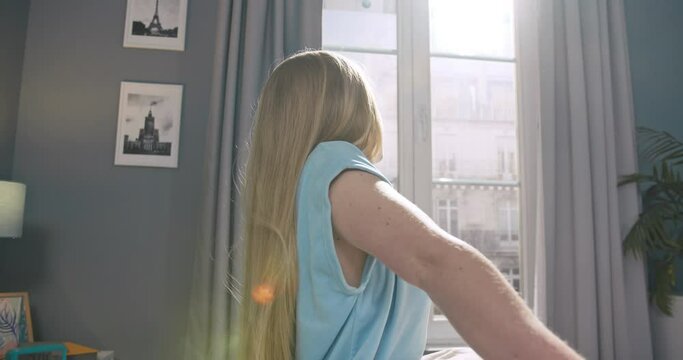 Beautiful young woman looking at the camera, sitting on bed, posing alone at home. Attractive female doing stratching and smiling to the camera in her apartment.