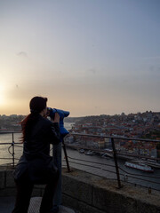 Fototapeta premium Young woman looks through coin binoculars to see Porto town in Portugal with Douro river. Traveler enjoying view panoramic point on vacation. Tourist at sunset visiting a scenic destination and using.