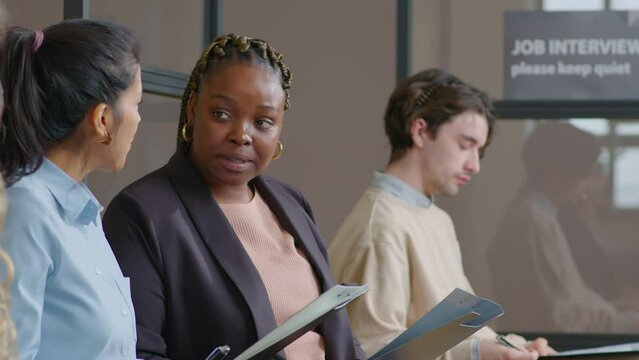 Tilt up shot of African American and Hispanic businesswomen discussing application form while sitting in line with other candidates and waiting for job interview