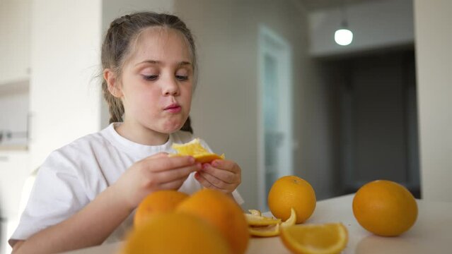 Little girl at kitchen table. Child eats yellow oranges. Happy child eats fruits. Vegetarian girl in kitchen.Concept of proper nutrition.Girl eats yellow fruit oranges.Juicy fruits proper healthy diet
