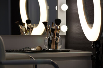 Mirror with light bulbs and ring lamp, near cosmetic brushes on dressing table indoors in makeup room. Shooting in a low key