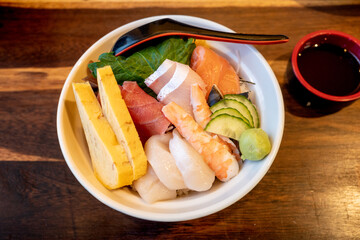 Top view of Sashimi Don, Danburi or Japanese rice bowl with various raw fresh slice of fish, shrimp, squid and tamago egg, on the wooden dark counter.