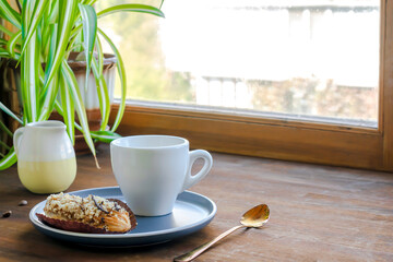 white espresso coffee cup, fresh baked eclair sweet dessert on plate kitchen table against window, utensils dishware, milk jug, home green plant. morning french home breakfast, copy space