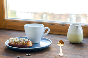 white espresso coffee cup, fresh baked eclair sweet dessert on plate kitchen table against window, utensils dishware, milk jug, home green plant. morning french home breakfast, copy space
